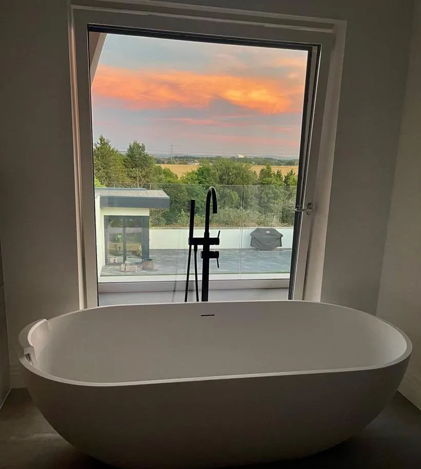 Freestanding bath and black floor-standing bath filler in front of a picture window at dusk