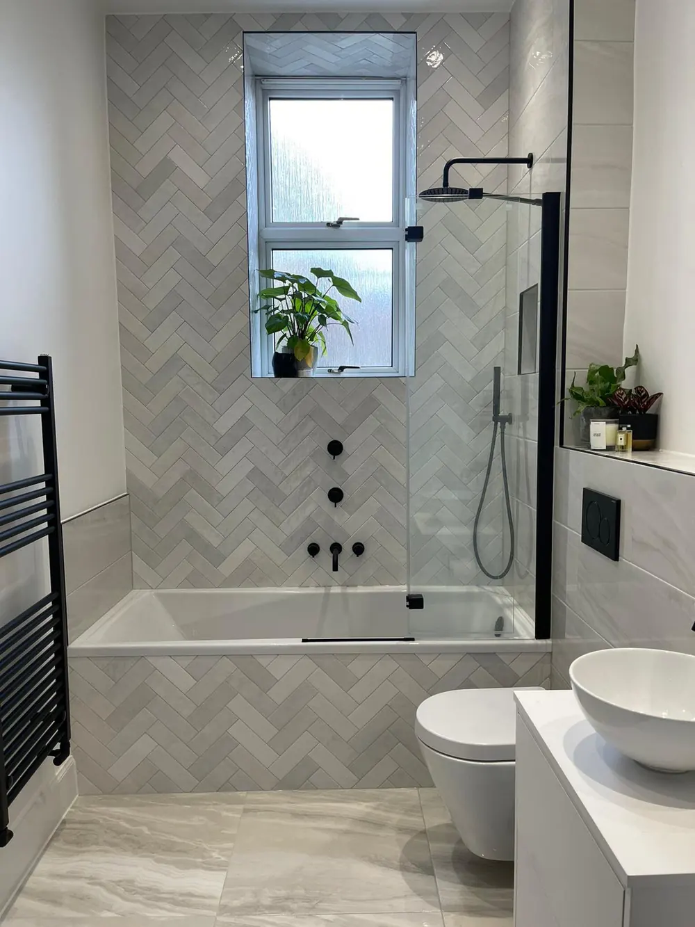 Bath with a shower over and a glass screen against a herringbone-tiled wall