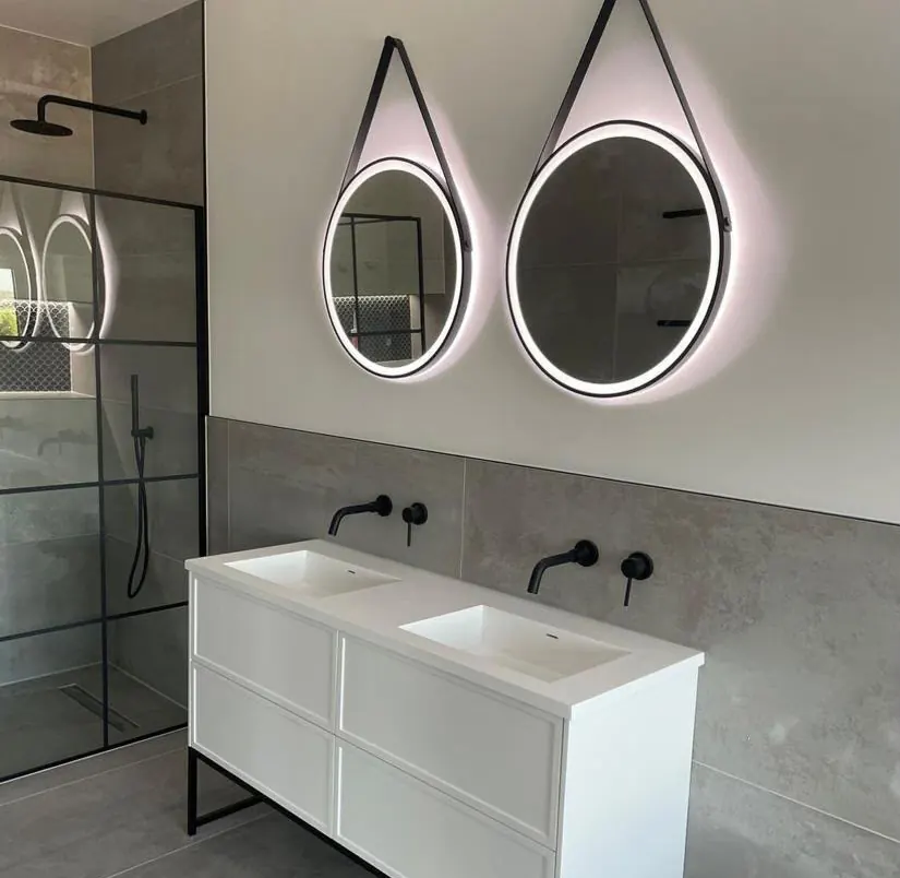 Twin basin vanity with backlit round mirrors beside a black-framed walk-in shower